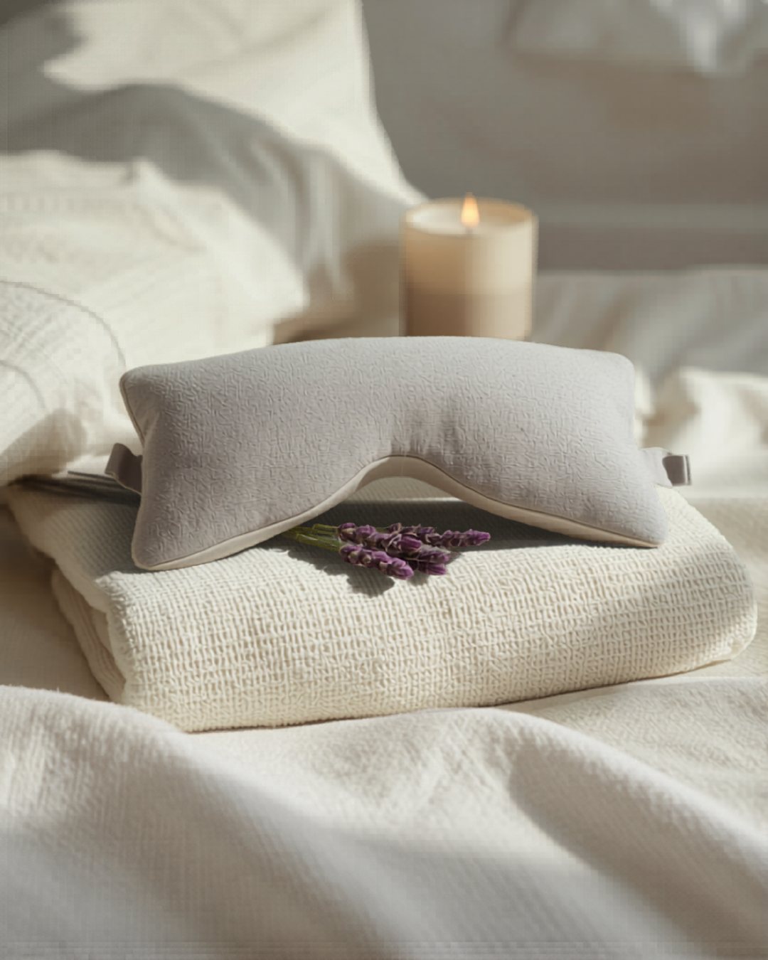 A lavender-grey weighted eye pillow on a cream waffle towel with fresh lavender and a soft candle